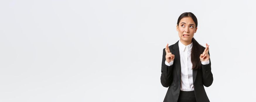 Hopeful asian businesswoman looking worried, biting lip and cross fingers good luck, making wish, anticipating big contract or deal, worried about outcome, standing white background
