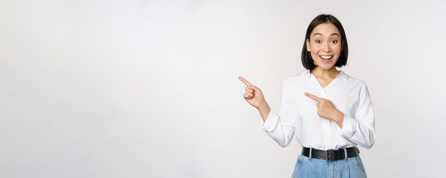 Portrait of excited young asian woman, office lady pointing fingers left at discount, showing sale banner, standing over white background