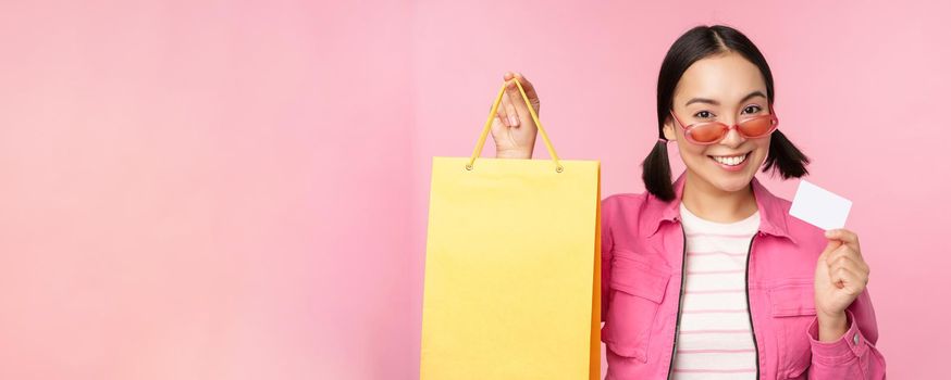 Happy young asian woman shows credit card and shopping bag, store sale announcement, buying smth in shop, posing against pink background