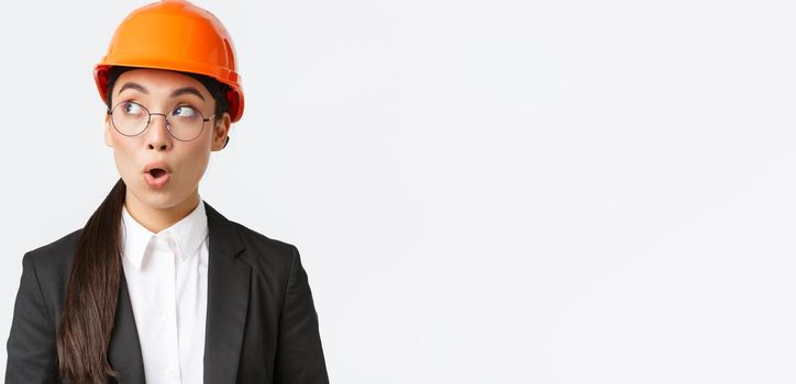Close-up of impressed asian female chief architect, industrial engineer in safety helmet and business suit, looking left surprised and amazed, standing white background at factory