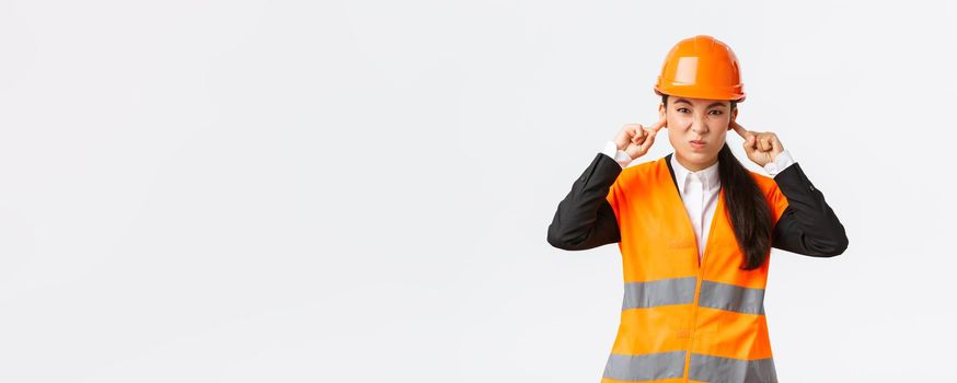 Bothered and displeased asian female chief engineer shut ears and grimacing from awful loud noise at construction area, wearing safety helmet, complaining on bothering sound, white background