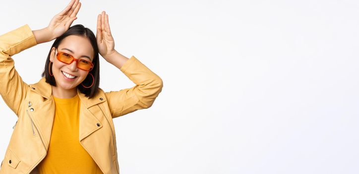 Portrait of beautiful asian girl in sunglasses, showing bunny ears hands gesture and smiling, standing happy over white background