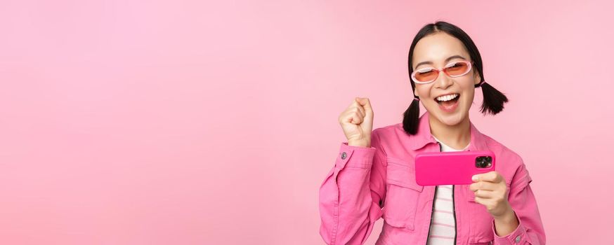 Happy smiling korean girl winning on mobile phone, looking at horizontal smartphone screen and rejoicing, achieve goal, celebrating, standing over pink background
