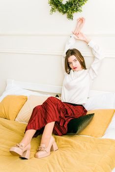 Portrait of fashionable women in red skirt, white blouse and stylish beige high-heeled shoes with a chain buckle posing in a bed
