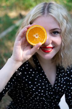 portrait of a charming blonde teenage girl with a fresh orange outdoors in forest or park. tasty juicy fruit