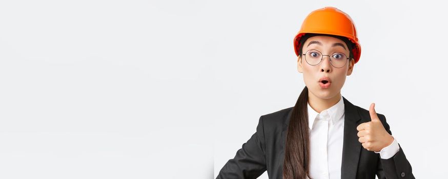 Close-up of professional pleased asian female chief engineer, architect in business suit and safety helmet showing thumbs-up in approval, give permission, recommend construction company