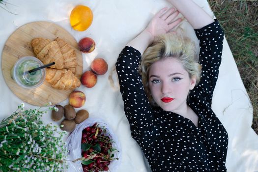 charming blonde young woman on a picnic on plaid in park with tasty snacks. Lemonade, fruits and croissants. summertime, rest, relax, enjoy. freedom