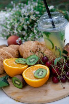 Healthy picnic for a summer day with croissants, fresh fruits and lemonade. peach, cherry, kiwi, oranges. white wildflowers