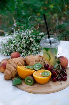 Healthy picnic for a summer day with croissants, fresh fruits and lemonade. peach, cherry, kiwi, oranges. white wildflowers