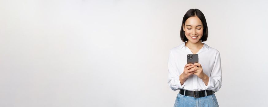 Stylish modern asian girl using mobile phone application, chatting on cellphone and smiling, standing in white blouse against studio background