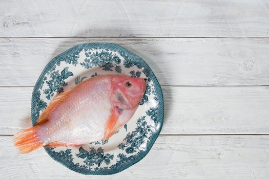 raw pink tilapia fish lies on a plate with blue ornaments on white wooden boards