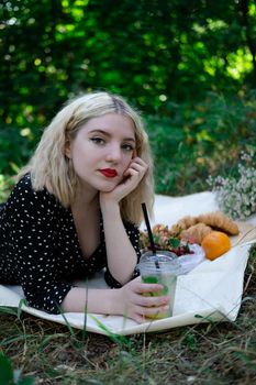 pretty blonde girl on picnic on white plaid in park with a plastic transparent cup with lemonade. fresh summer refreshing drink