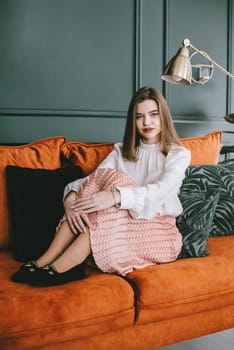 Portrait of fashionable young woman in a beige skirt, white blouse and stylish beige black seude shoes with a buckle posing on a orange sofa.