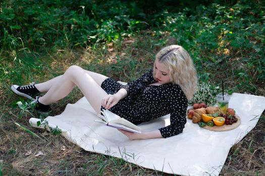portrait of young woman on a picnic on plaid in park reading a book with tasty snacks