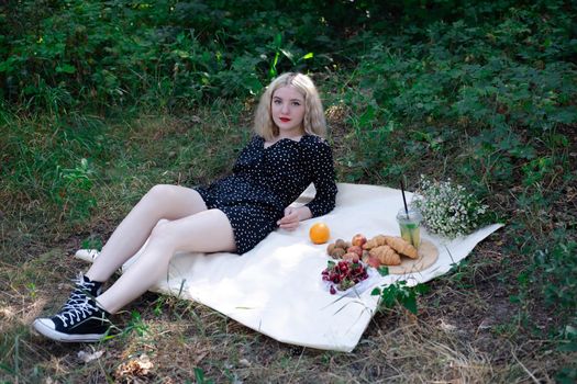 pretty blonde girl on a picnic in a forest or park with lemonade, fruits and croissants. summertime, rest, relaxation. millennial generation