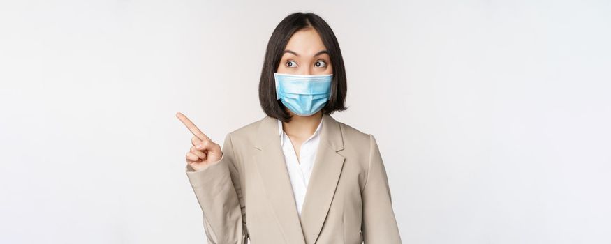 Enthusiastic businesswoman pointing fingers left, wearing medical face mask from covid-19 pandemic, standing over white background
