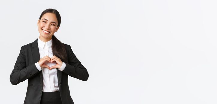 Cheerful successful asian businesswoman in black suit care for her clients, showing heart gesture and smiling happy, providing caring service for customers, standing white background