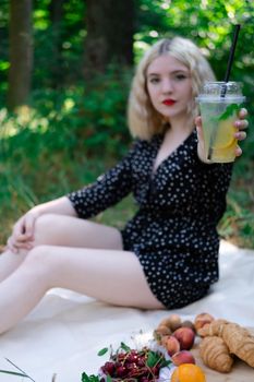 pretty blonde girl on picnic on white plaid in park with a plastic transparent cup with lemonade. fresh summer refreshing drink