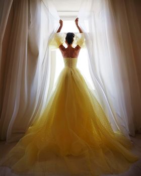 A woman's silhouette in a golden luxurious dress against the background of a window holds a curtain with her hands. Elegant lady in a yellow long silk dress with bare back, back view.