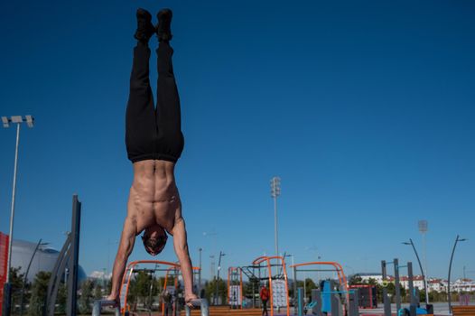 Shirtless man doing handstand on parallel bars at sports ground.