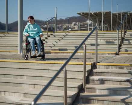 A caucasian woman in a wheelchair cannot go down stairs without a ramp. Hard to access environment.