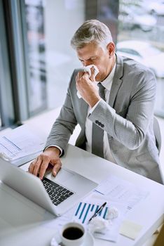 It is not productive to be sick at work. Cropped shot of an ill businessman blowing his nose in the office while working on a laptop.