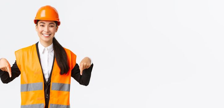 Smiling cheerful construction manager, asian female engineer in safety helmet and reflective clothing showing announcement. Woman project manager pointing down, standing white background