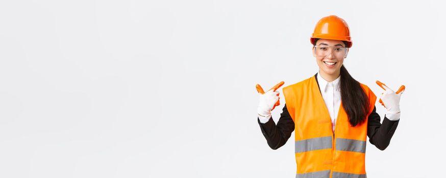 Smiling confident asian female chief construction engineer in safety helmet, gloves and glasses pointing at herself, notify to follow safe protocol and wearing special clothing, white background