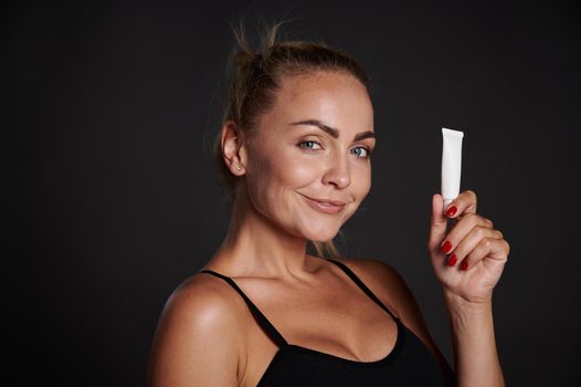 Beautiful woman with tanned skin holding a tube of anti wrinkles smoothing anti puffiness under eyes cream, smiles looking at camera, isolated over black background with copy ad space