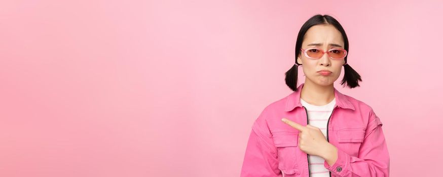 Potrait of cute korean girl in sunglasses, pointing left and looking disappointed, sulking upset, standing over pink background