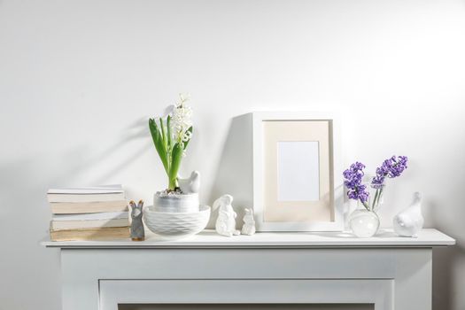 White hyacinth in a large porcelain bowl, books, figurines of hares and a bird, an empty photo frame are on the fireplace against the white wall. Layout.