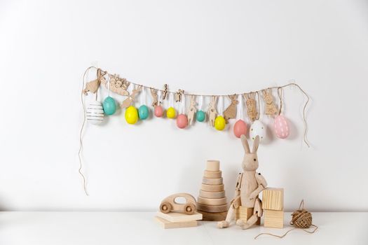 Fragment of the interior. Decorated children's room for Easter. A garland of plastic eggs and hares cut out of cardboard on the wall. Wooden rabbits and wooden cubes on the table. Place for your text. Easter card.
