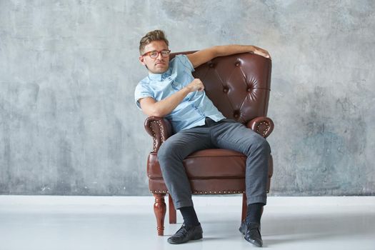 Portrait of a stylish intelligent man stares into the camera, small unshaven, charismatic, blue shirt, sitting on a brown leather chair, dialog, negotiation, short sleeve, brutal, hairstyle