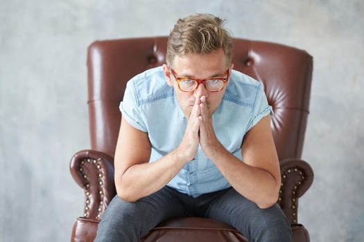 Portrait of a stylish man with glasses, stares into the camera, unshaven, charismatic, blue shirt with short sleeve, sitting on a brown leather chair, dialogue, negotiation, hands as in prayer