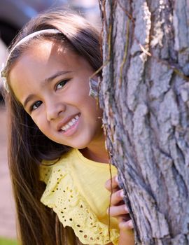 Dont tell anyone about my favourite hide-and-seek spot. Shot of an adorable little girl hiding behind a tree.