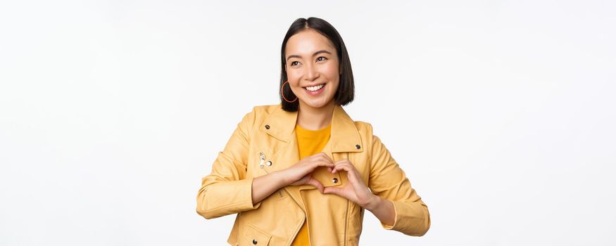 Image of beautiful asian girl showing heart, love sign, express care and romantic feelings, standing over white studio background
