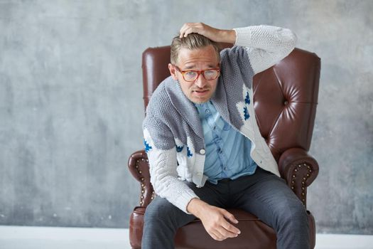 Portrait of a stylish intelligent man with glasses stares into the camera, good view, small unshaven, charismatic, blue shirt, gray sweater, sitting on a brown leather chair, grabs his head