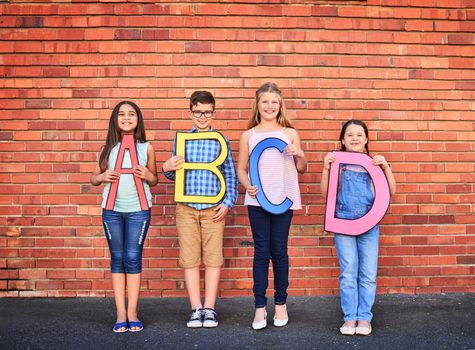 The more you know, the more you grow. Portrait of a group of young children holding letters from the alphabet against a brick wall.