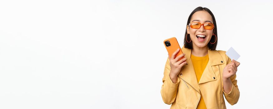 Online shopping and delivery concept. Happy korean girl in stylish clothes, holding credit card and smartphone, laughing and smiling, standing over white background