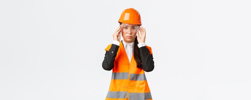 Serious-looking young asian female construction manager follow safety protocol, wearing protective glasses and helmet before entering enterprise, standing white background