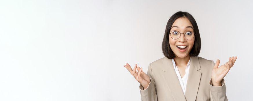Enthusiastic asian saleswoman in glasses, smiling and laughing, looking amazed at camera, standing in beige suit over white background