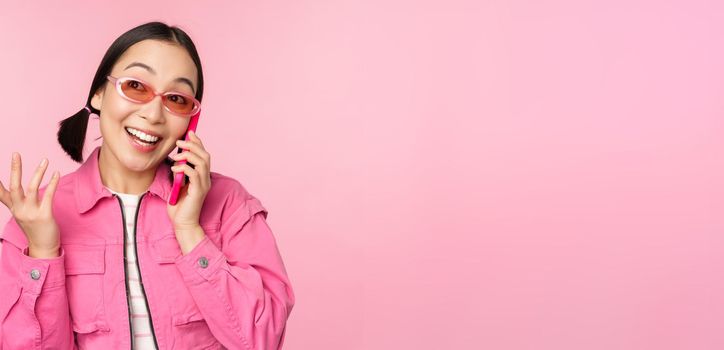 Beautiful korean female model in sunglasses, talking on mobile phone with happy face, using cellular service to call friend on smartphone, standing over pink background