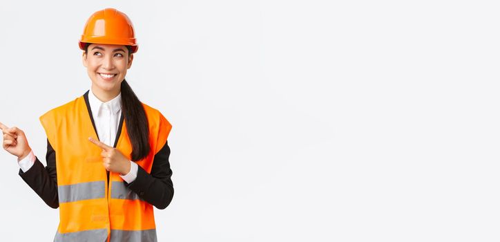 Building, construction and industrial concept. Smiling confident asian female chief engineer in safety helmet and reflective clothing pointing fingers upper left corner and looking satisfied
