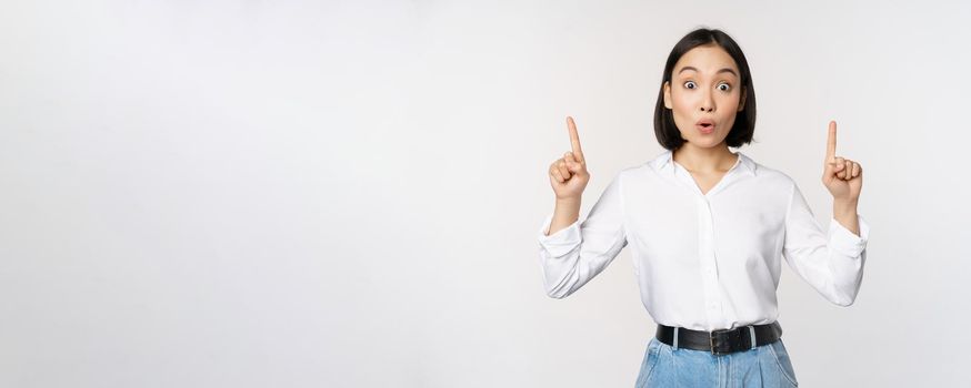 Surprised asian girl, pointing fingers up, showing top banner, information or advertisement, standing over white background in blouse