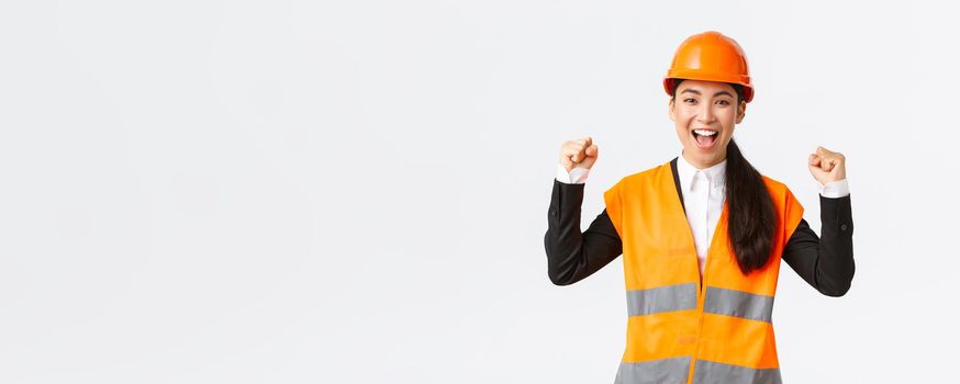 Successful winning female asian engineer fist pump and shout yes delighted, wear safety helmet and jacket, triumphing over victory, finish building in time, standing satisfied white background