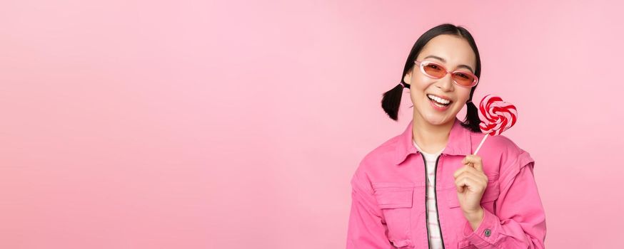 Stylish korean girl licking lolipop, eating candy and smiling, standing in sunglasses against pink background