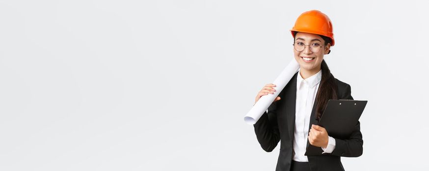 Successful asian female architect in business suit and helmet holding blueprints and clipboard with notes, inspector looking at construction works, smiling at camera, white background