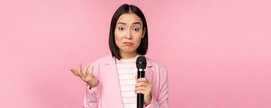Indecisive, nervous asian business woman holding mic, shrugging and looking clueless, standing with microphone against pink background, wearing suit