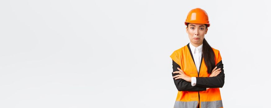 Awkward asian female engineer in reflective jacket, safety helmet, cross arms chest and smirk surprised, making silly mistake, acting natural, standing white background