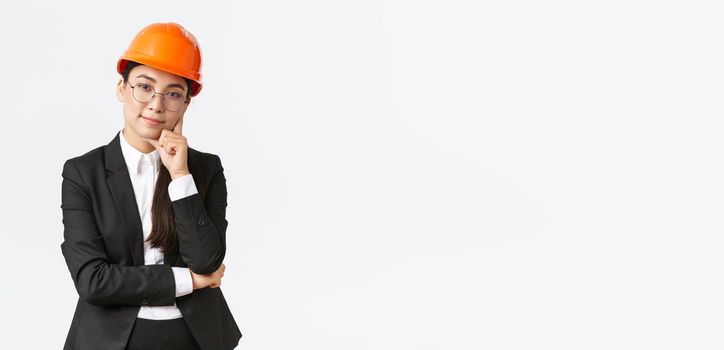 Smiling professional asian businesswoman invest money in construction, manage work at enterprise, wearing safety helmet and suit, looking pleased at camera, standing white background